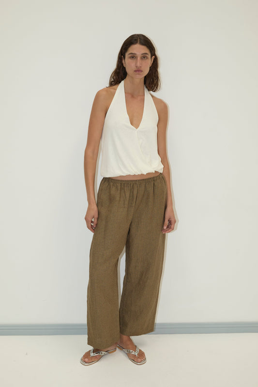A woman with long brown hair and a neutral expression stands against a white wall, wearing loose brown Ease Trousers, a sleeveless white halter top, and flip-flops.