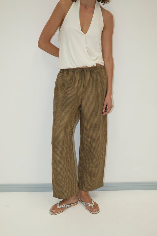 A woman stands against a plain background wearing a sleeveless white wrap top, flip-flops, and the Linen Ease Trouser in Pine Check. Her right hand rests on her hip, and her head is not visible in the image.