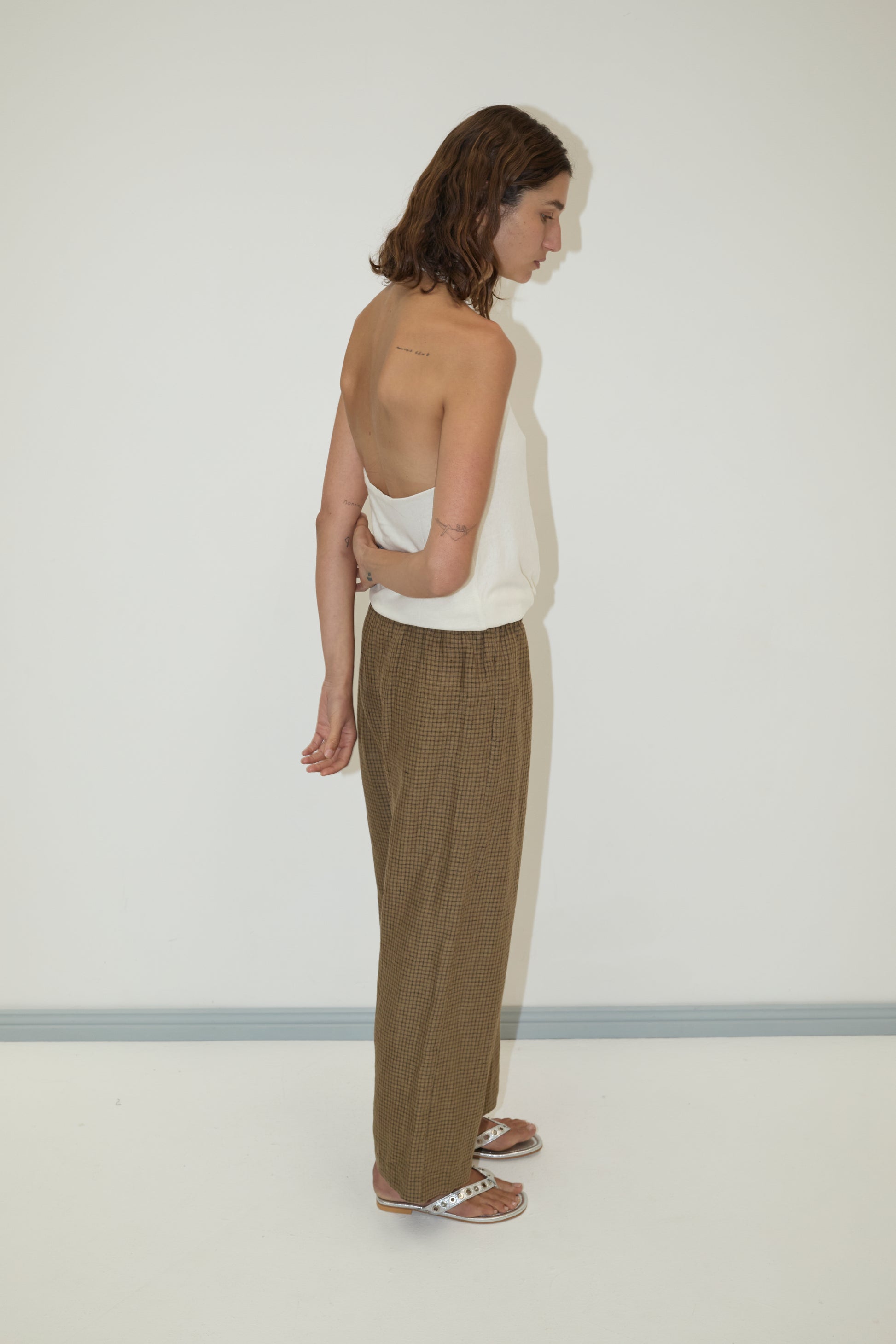 A woman with shoulder-length brown hair stands sideways against a white wall, wearing a white sleeveless top, sandals, and the Linen Ease Trouser - Pine Check. Her left arm rests across her back.