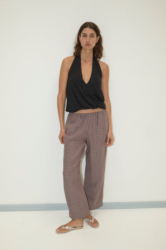 A woman with long brown hair and a neutral expression stands against a white wall wearing a sleeveless black halter top, flip-flops, and Deiji Studios linen ease trouser in russet check.