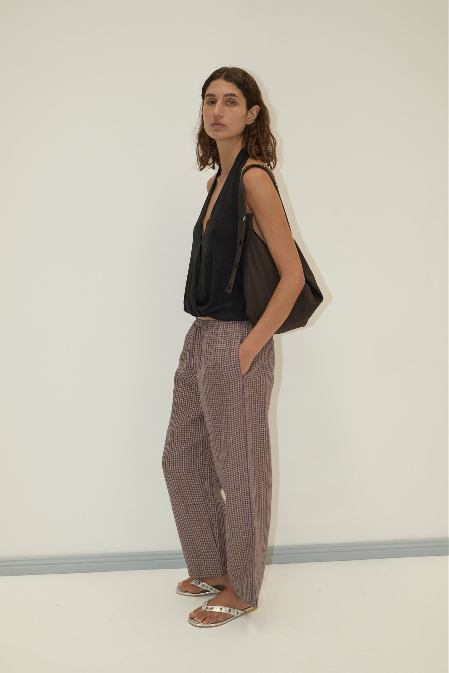 A woman stands against a white wall, wearing a loose black sleeveless top and Deiji Studios’ linen ease trouser - russet check in soft French linen. She pairs them with sandals, a large dark bag over her shoulder, and hands in her pockets.