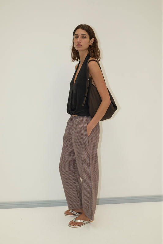 A woman stands against a white wall, wearing a loose black sleeveless top and Deiji Studios’ linen ease trouser - russet check in soft French linen. She pairs them with sandals, a large dark bag over her shoulder, and hands in her pockets.