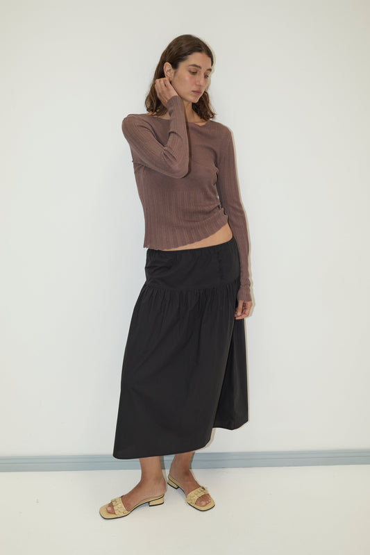 A woman with long brown hair stands against a plain wall wearing a long-sleeved brown top, the Deiji Studios Button Skirt in Black made from organic cotton, and beige sandals, looking down as she gently touches her neck.