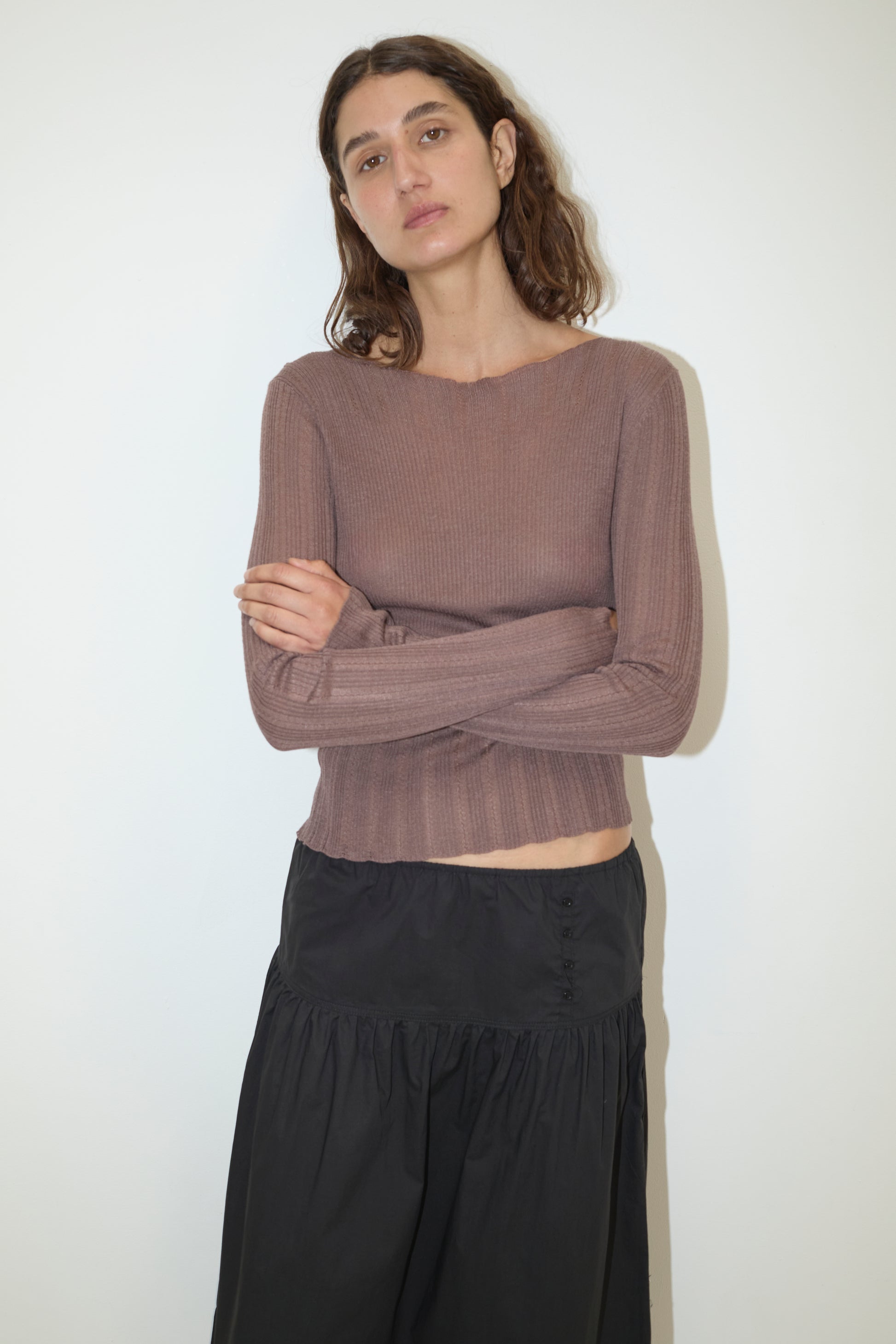 A woman with wavy brown hair stands against a white wall, wearing a long-sleeved, ribbed brown top and the Button Skirt - Black from Deiji Studios. She has her arms crossed and a neutral expression.