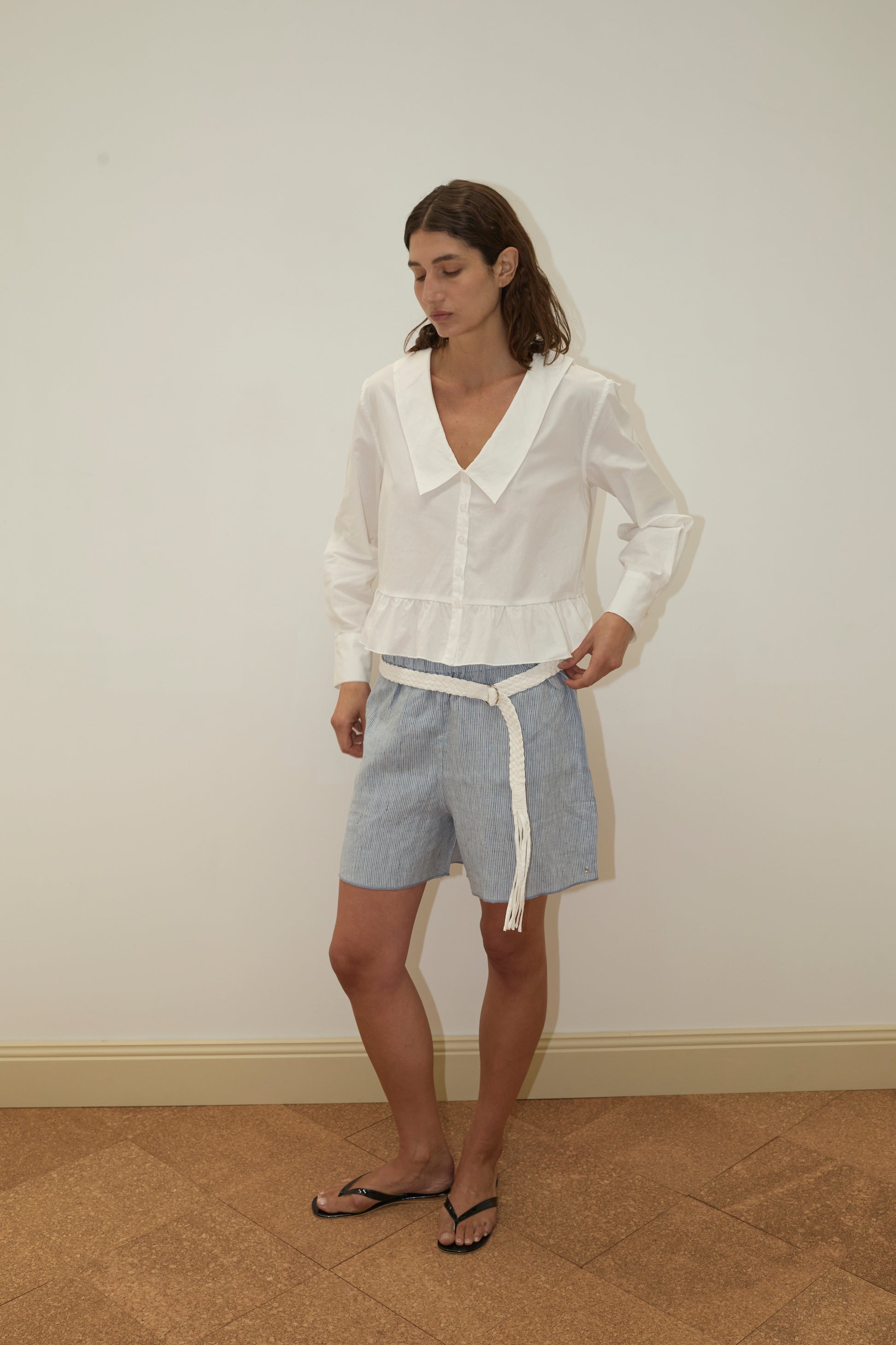 Wearing the Sweep Shirt in white organic cotton poplin and blue striped shorts with a white rope belt, a person stands indoors on a patterned brown floor against a plain light wall, looking slightly downward.
