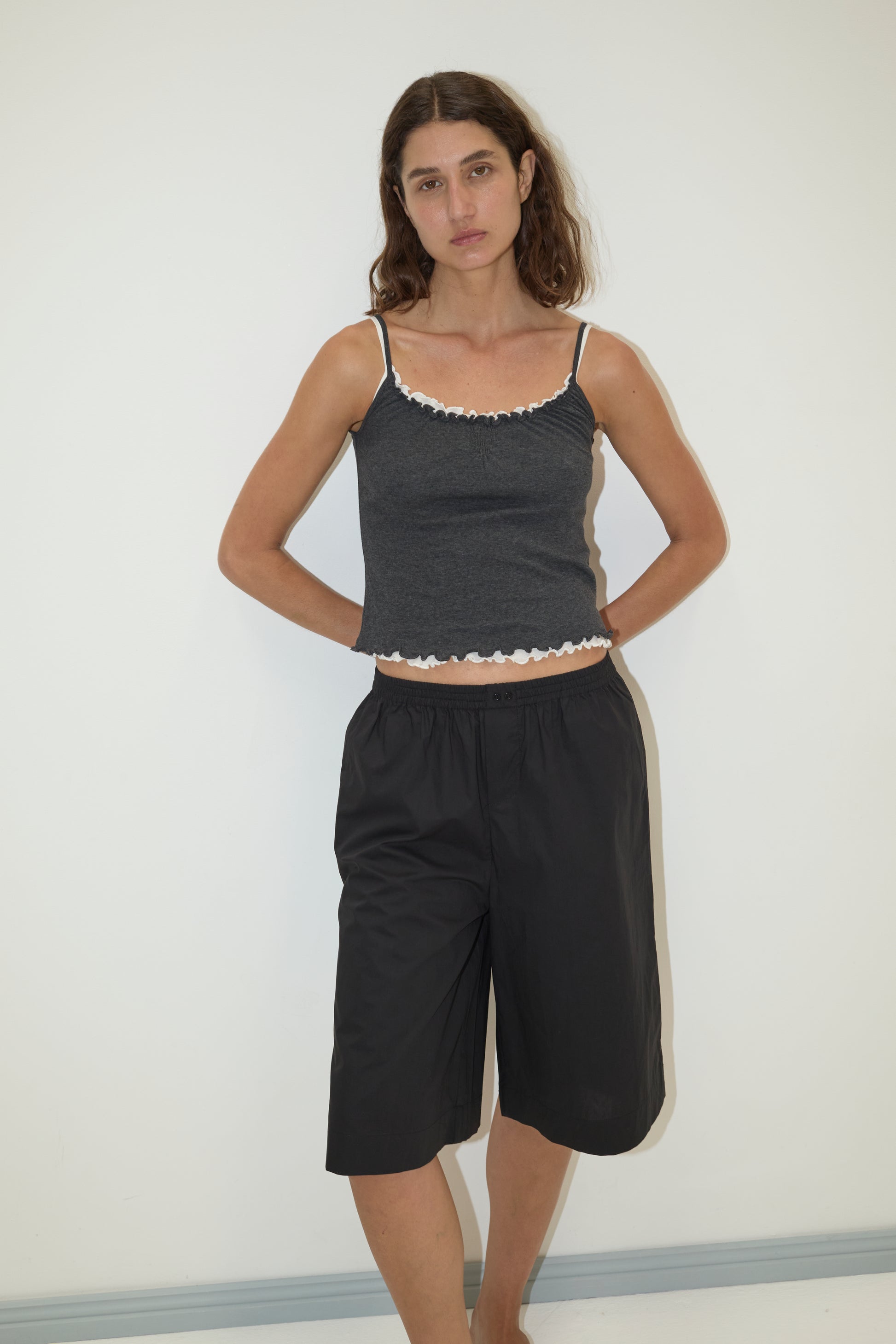 A woman with long brown hair stands against a white wall, wearing the Long Lounger - Black by Deiji Studios. The relaxed knee-length piece is crafted from organic cotton and features a dark hue. She has a neutral expression, hands clasped behind her back.