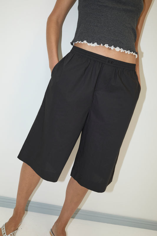 A person wearing the Deiji Studios Long Lounger in black, a dark gray sleeveless top with a ruffled hem and loose knee-length shorts, stands against a white wall with hands in pockets.