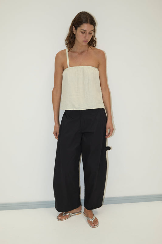 A woman stands against a plain white wall wearing a cream asymmetrical strap top, the black Tab Pant from Deiji Studios, and silver sandals. She looks slightly downward with her arms relaxed at her sides.