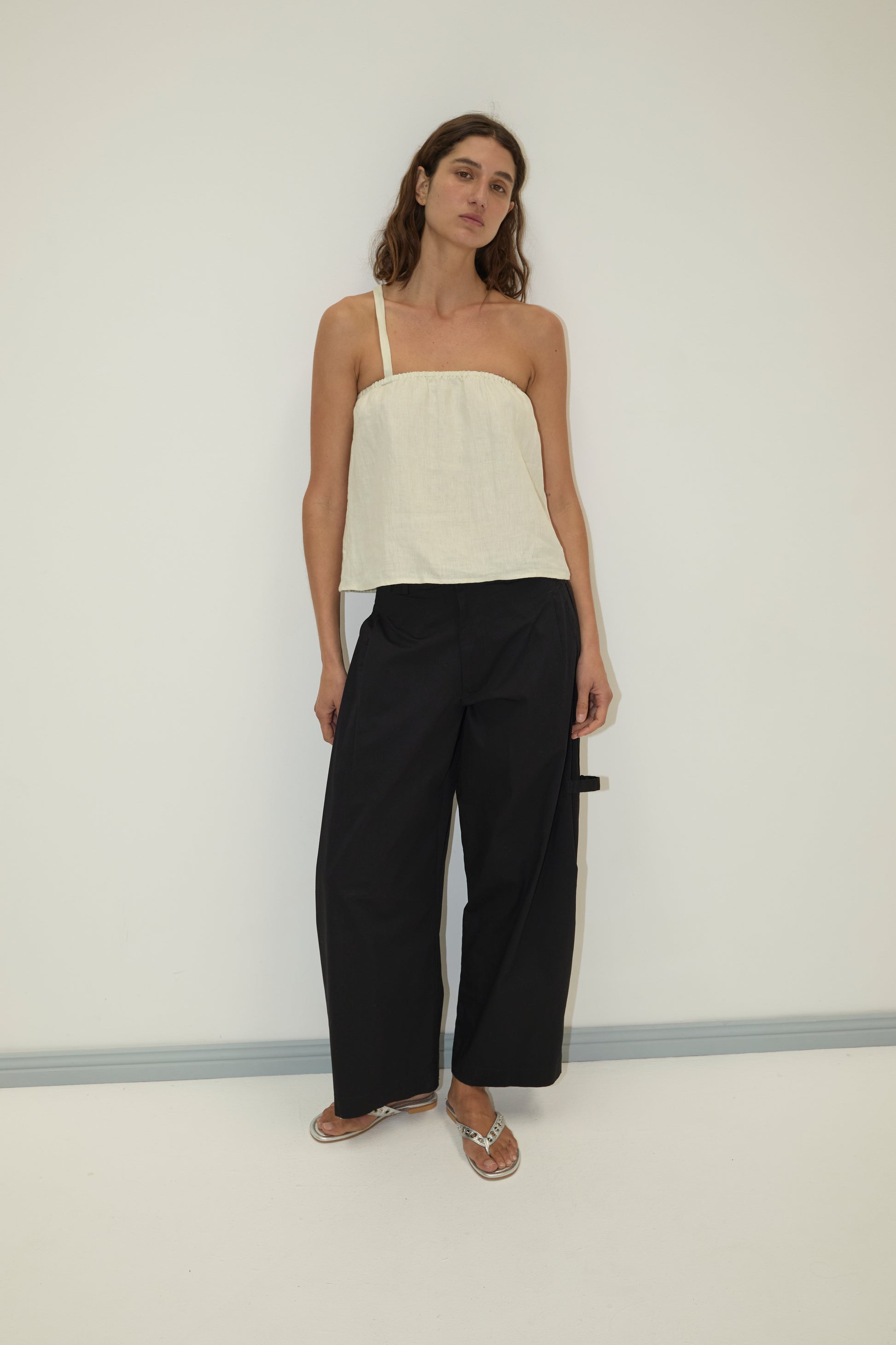 A woman stands against a plain wall wearing the Button Strap Top - Pastel Pistachio from Deiji Studios, crafted from organic French linen. She pairs it with loose black wide-leg pants and silver sandals, standing relaxed with one arm slightly bent.