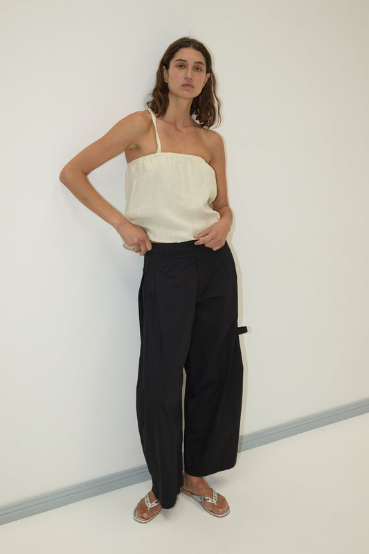 A woman stands against a white wall, wearing a cream one-shoulder top, the Tab Pant - Black from Deiji Studios in organic cotton, silver flip-flops, and has long wavy brown hair with a relaxed, confident pose.