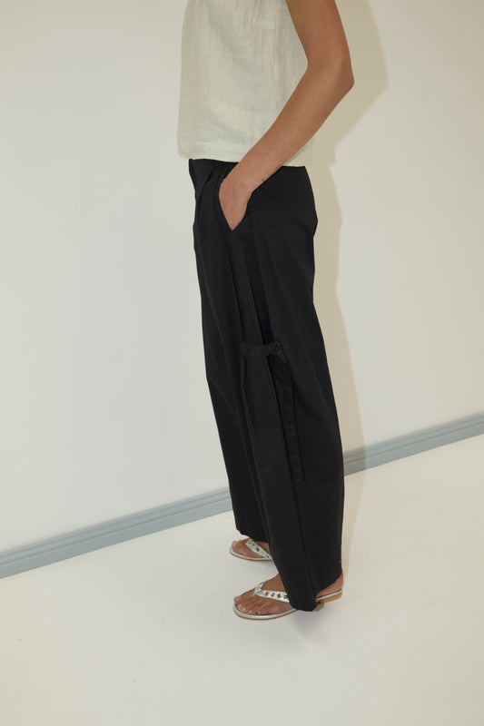 A person stands sideways with one hand in their pocket, wearing a sleeveless light beige top, Tab Pant - Black by Deiji Studios, and white sandals against a plain light background.