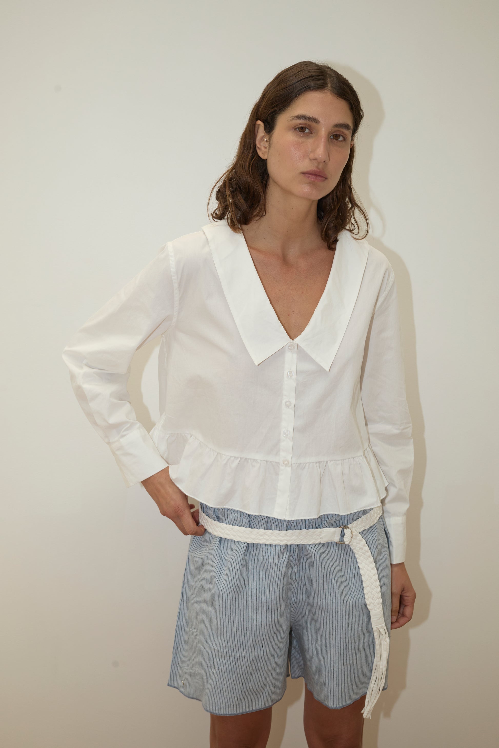 A woman with wavy brown hair and a neutral expression stands against a plain background wearing the Deiji Studios Sweep Shirt in white, featuring a wide collar and ruffled hem, styled with light blue striped shorts and a white braided belt.