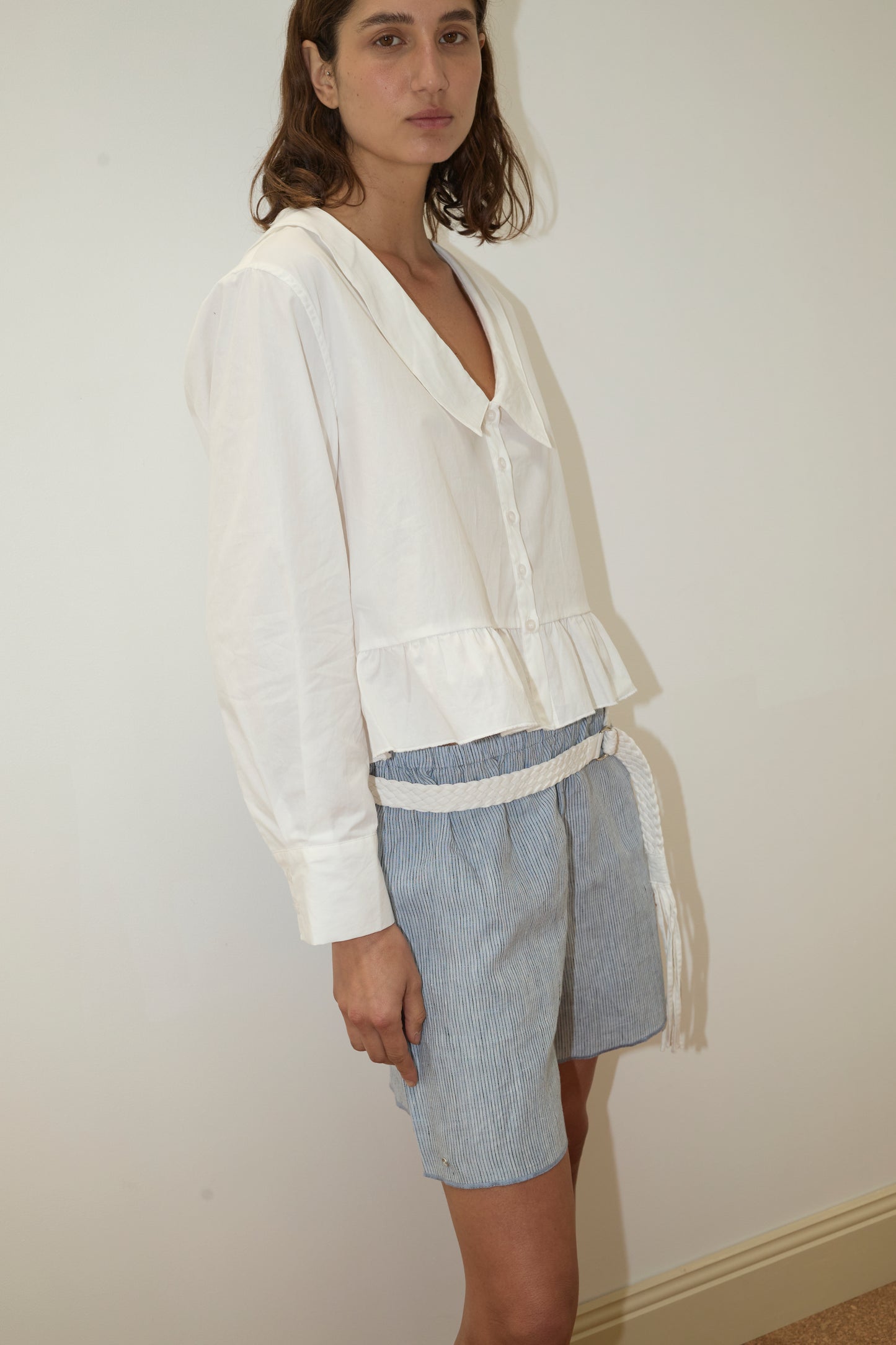 A woman stands against a plain wall, wearing the Sweep Shirt - White by Deiji Studios—a loose organic cotton poplin blouse with long sleeves and a ruffled hem—paired with blue and white striped shorts and a white braided belt.