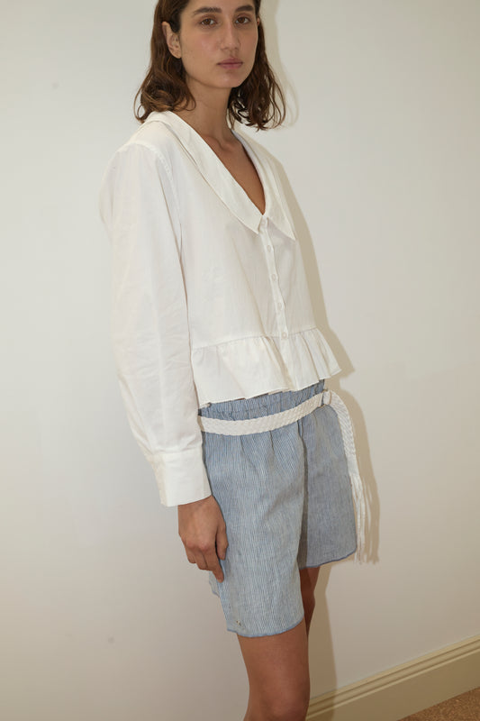 A woman stands against a plain wall, wearing the Sweep Shirt - White by Deiji Studios—a loose organic cotton poplin blouse with long sleeves and a ruffled hem—paired with blue and white striped shorts and a white braided belt.