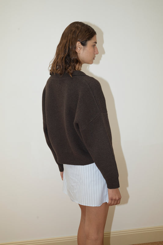 A person with wavy brown hair stands sideways, wearing the Deiji Studios Collared Pullover in Burnt Wood and a short, light blue pinstriped skirt, against a plain light-colored wall.