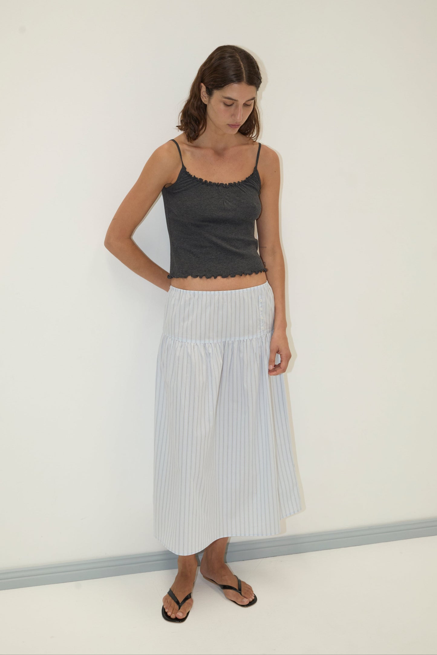 A woman stands against a plain white wall, wearing a dark gray sleeveless top, the Button Skirt—a light-colored striped midi skirt—and black flip-flops. She looks down with her arms behind her back.