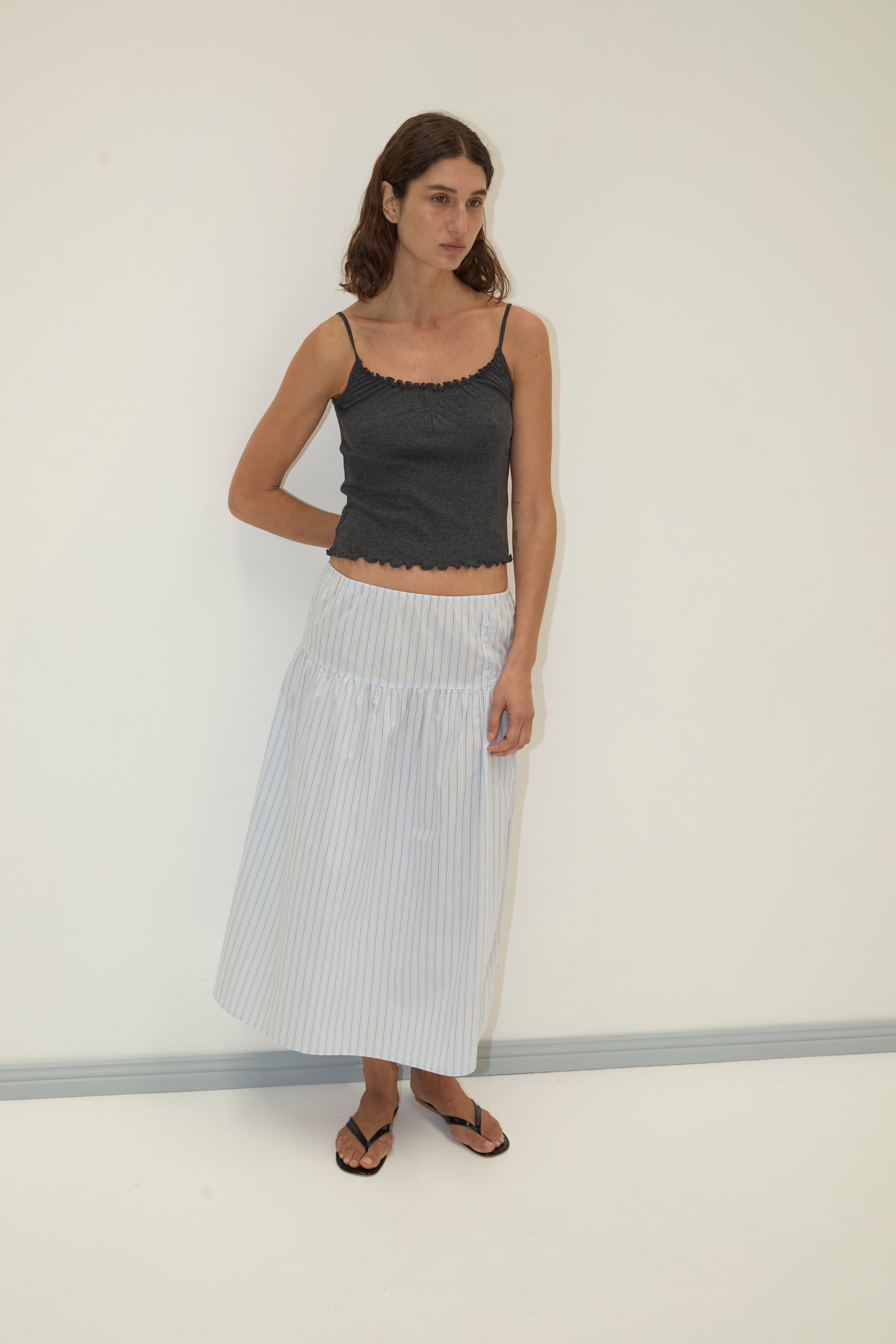 A woman with long brown hair wears a dark gray camisole, black sandals, and the Button Skirt - Mineral Stripe, an ankle-length white organic cotton skirt with a subtle pattern. She stands against a plain white wall, looking slightly to the side.