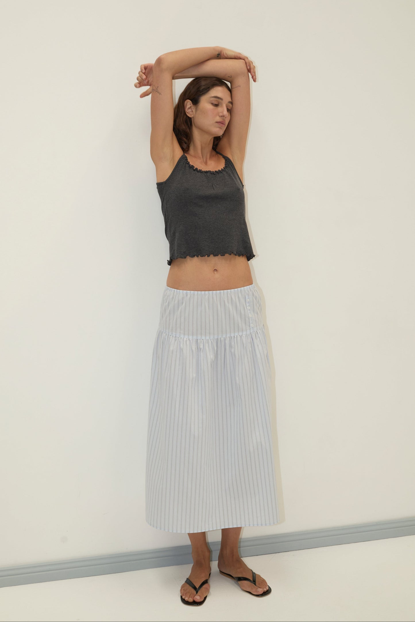 A woman stands against a white wall with arms raised and eyes closed, wearing a dark gray sleeveless crop top, the Button Skirt - Mineral Stripe from Deiji Studios in light blue and white stripes, and black sandals.