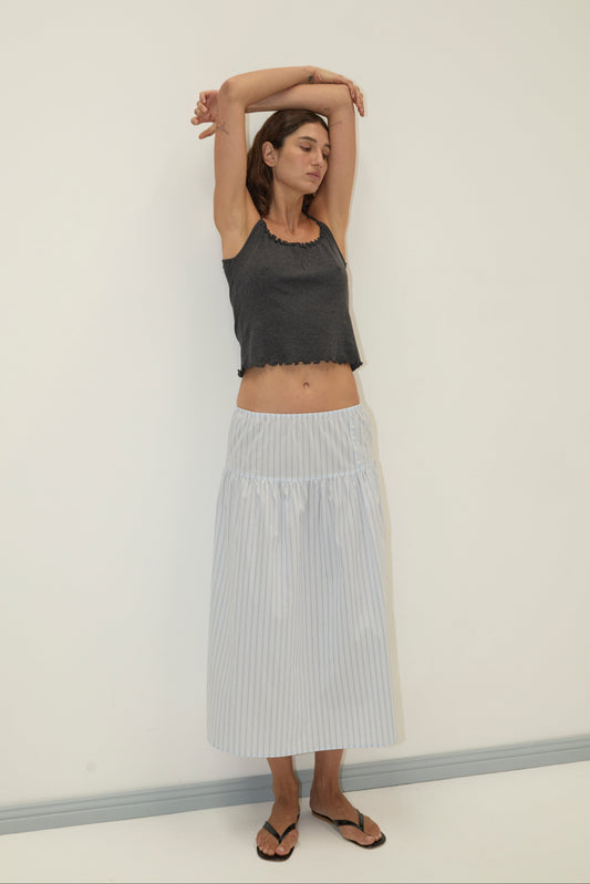 A woman stands against a white wall with arms raised and eyes closed, wearing a dark gray sleeveless crop top, the Button Skirt - Mineral Stripe from Deiji Studios in light blue and white stripes, and black sandals.