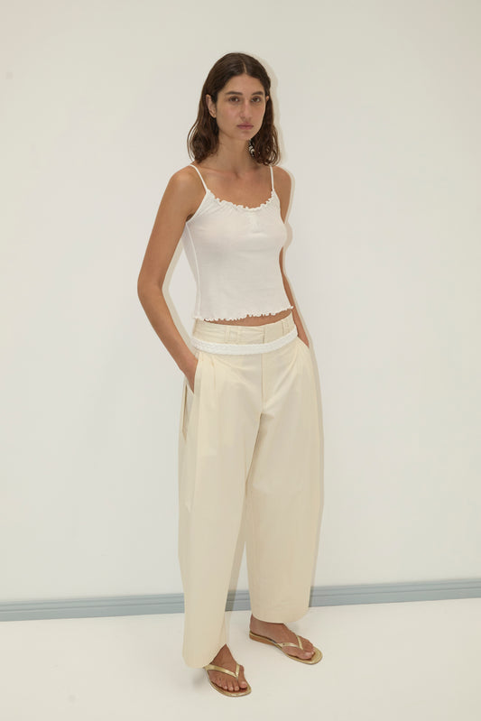 A woman stands against a plain white wall, wearing a fitted white tank top and Deiji Studios' Tab Pant in Off White—high-waisted, wide-leg trousers with her hands in her pockets and beige sandals.
