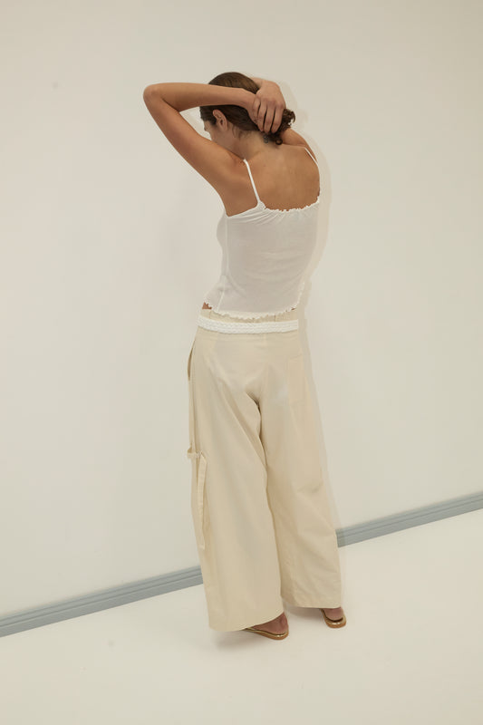 A person stands with their back to the camera, arms raised and hands behind their head. They wear a white tank top and the Tab Pant - Off White by Deiji Studios in a minimal, light-colored room.