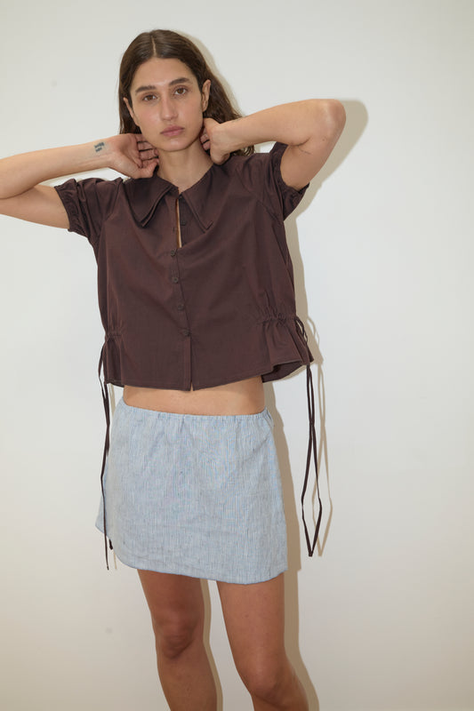 Against a plain white background, a person wears the Deiji Studios Double Collar Shirt - Umber, paired with a light blue mini skirt. They adjust their hair and look at the camera with a neutral expression.