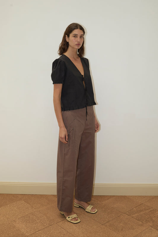A woman with shoulder-length brown hair stands indoors against a light wall, wearing a black short-sleeve top, Deiji Studios Adjust Pocket Pant - Sawdust in brown organic cotton, and open-toe sandals. She has a neutral expression.