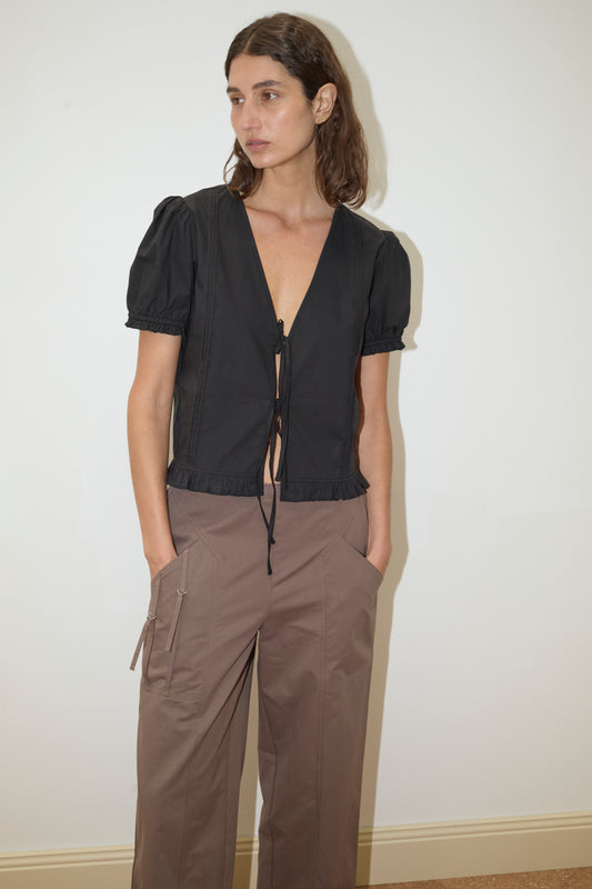 A person with long brown hair stands against a white wall, wearing a Deiji Studios Pintuck Top in black with short sleeves and a deep V-neck, paired with brown cargo pants, hands in pockets, looking slightly to the side.
