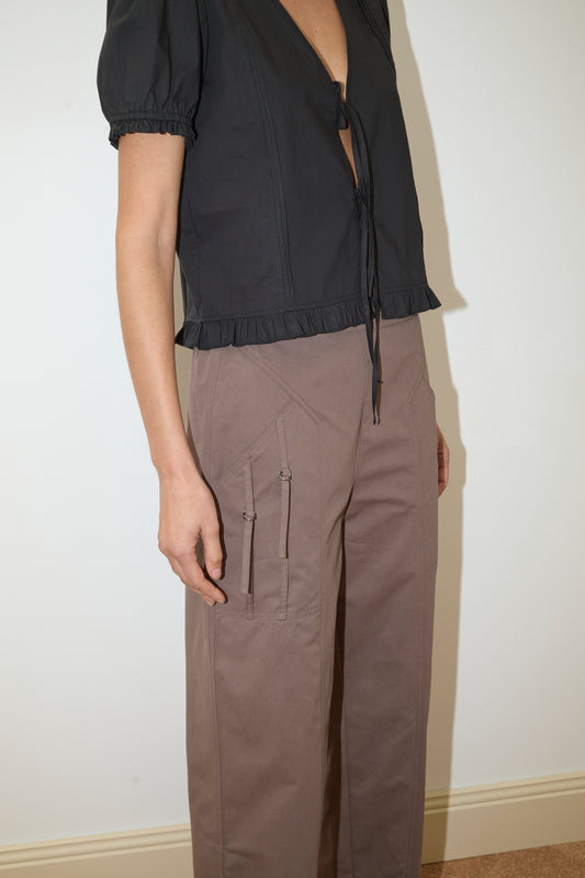 A person wears the Deiji Studios Pintuck Top - Black, a short-sleeved organic cotton poplin top with a deep V-neck, chest tie, and ruffle details at the hem and sleeves, paired with taupe wide-leg pants against a plain light wall.