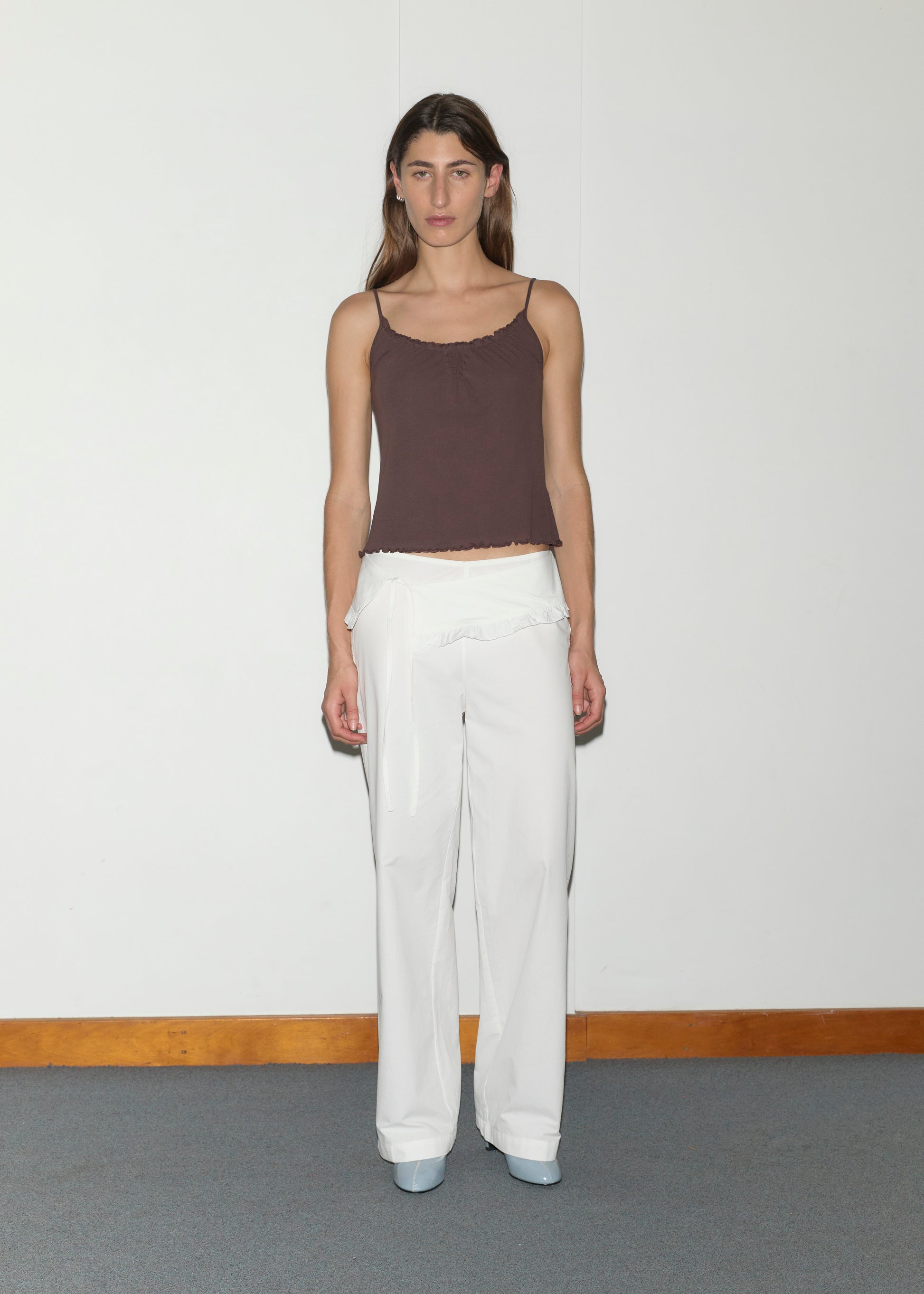 A woman with straight brown hair and a neutral expression stands against a white wall, wearing Deiji Studios' Cotton Flounce Tank in brown, white wide-leg ruffled pants, and light blue platform shoes.
