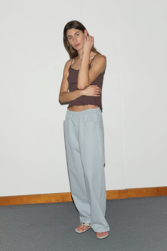A person stands indoors against a white wall, wearing a brown sleeveless top, Square Pocket Pant - Blue by Deiji Studios, and sandals. They have long brown hair and are posing with one arm crossed and the other touching their neck.