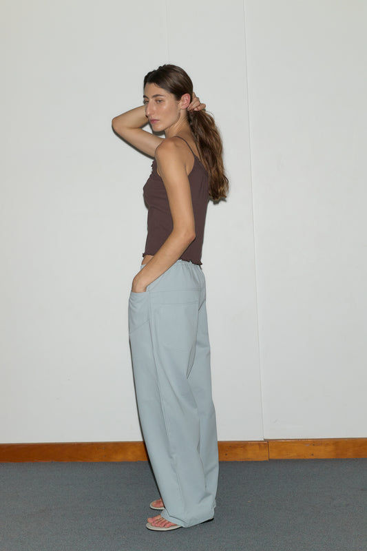A woman stands sideways against a plain white wall, wearing a brown sleeveless top and Deiji Studios' Square Pocket Pant - Blue. She appears relaxed in sandals with one hand raised to her hair.