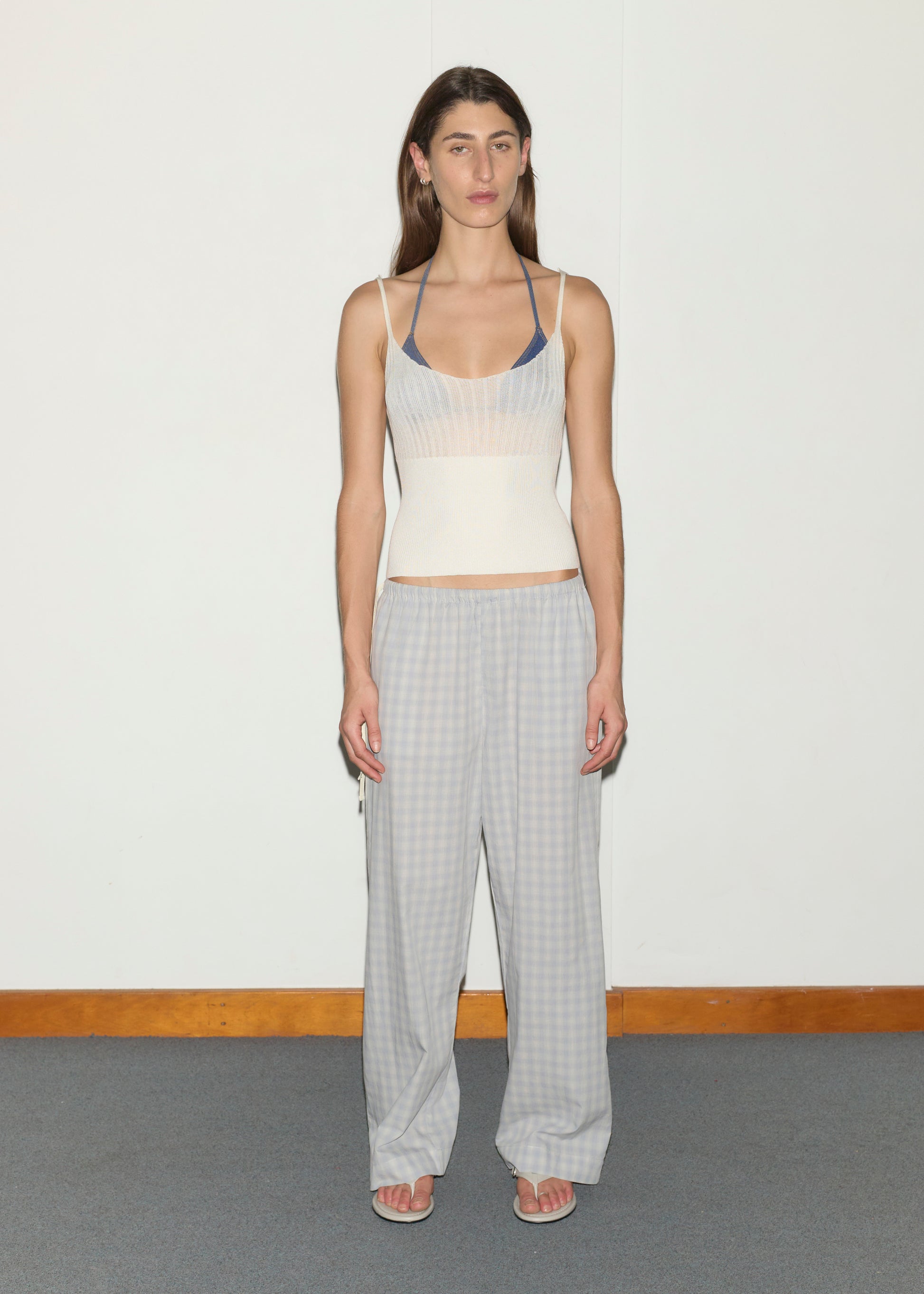 A person stands indoors against a plain white wall, wearing a cream fitted tank over a blue bralette, Side Tie Pant - Blue Check, and white flip-flops. Their expression is neutral with arms relaxed at their sides.