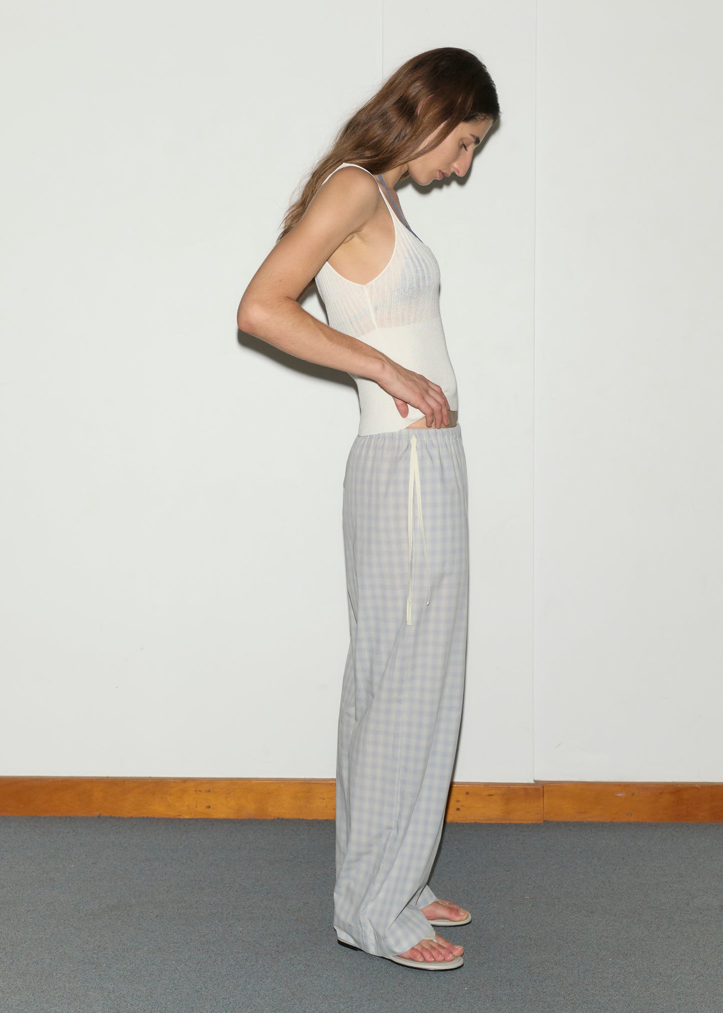 A woman stands sideways against a plain wall, wearing a white tank top, the Side Tie Pant - Blue Check in soft cotton poplin, and white sandals. She looks down with one hand resting on her waist.