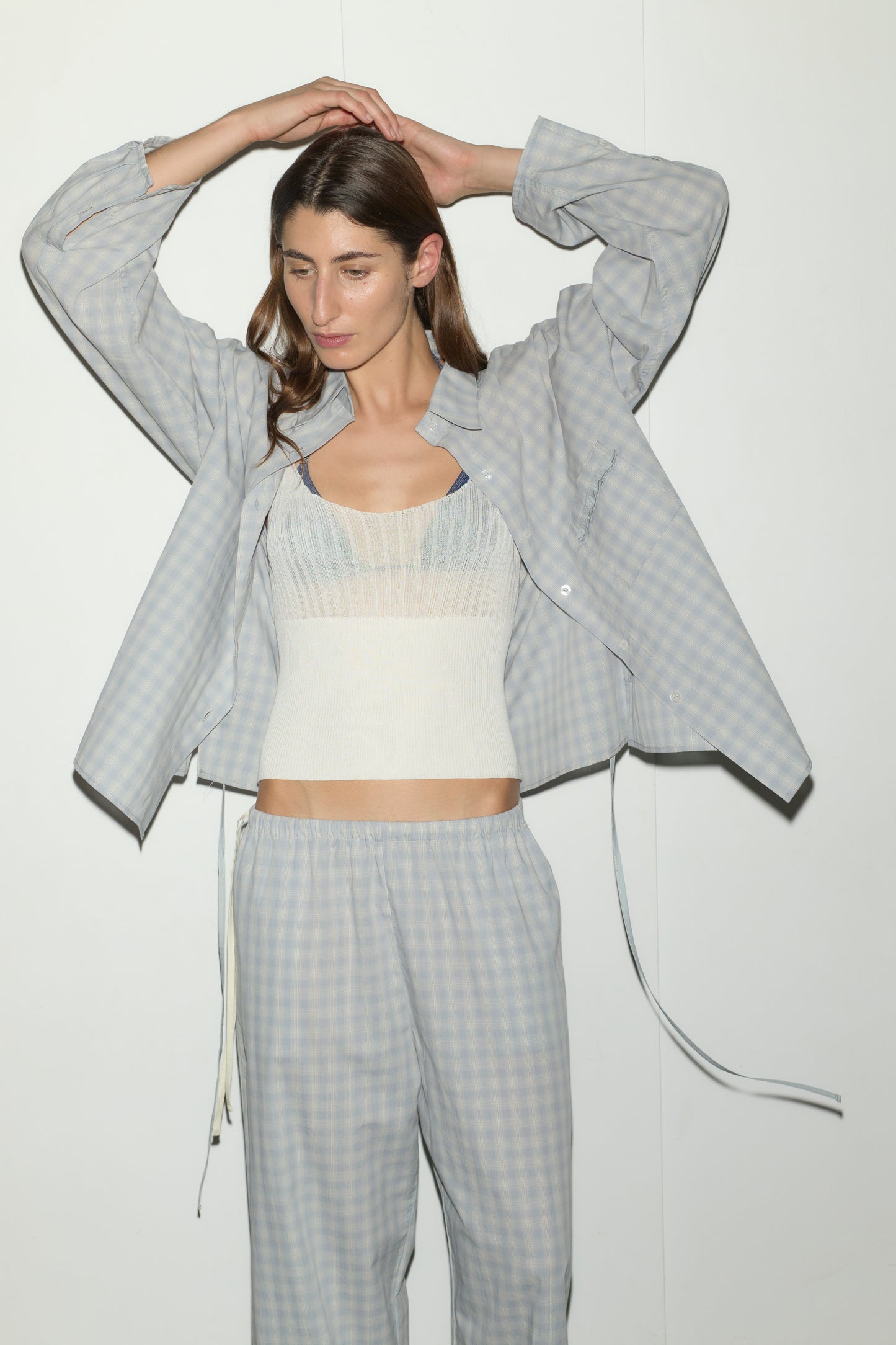 A woman stands against a white wall, wearing the Side Tie Pant - Blue Check with a matching open shirt over a white tank. Her arms are raised and her long hair is loose.