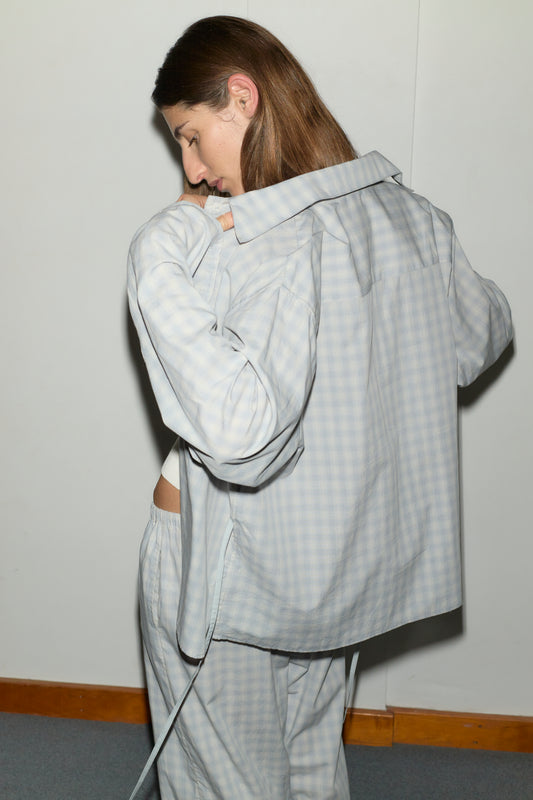 A person with long brown hair stands sideways, looking down and adjusting the collar of the Day Shirt - Blue Check. The shirt has a boxy fit and light blue check pattern. Part of their side is visible against a plain background with a wooden trim.