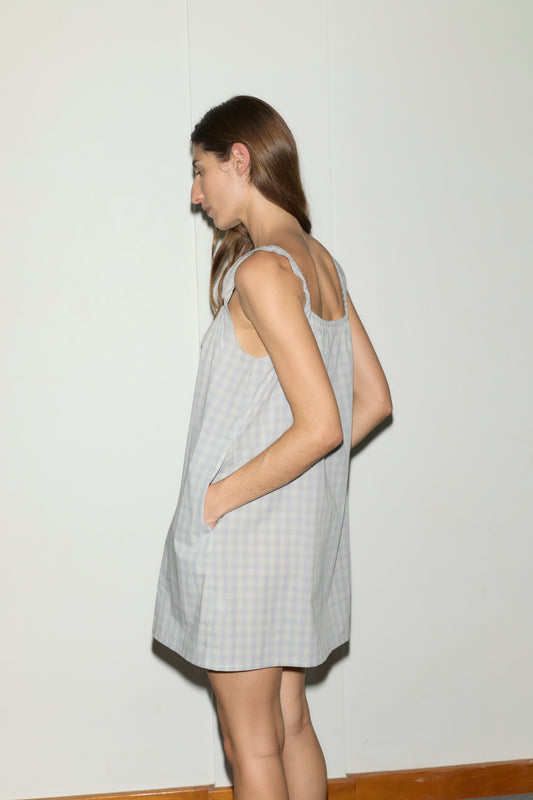 A person with long brown hair stands sideways against a plain white wall, wearing the bow dress - blue check by Deiji Studios, a loose sleeveless style with pockets.