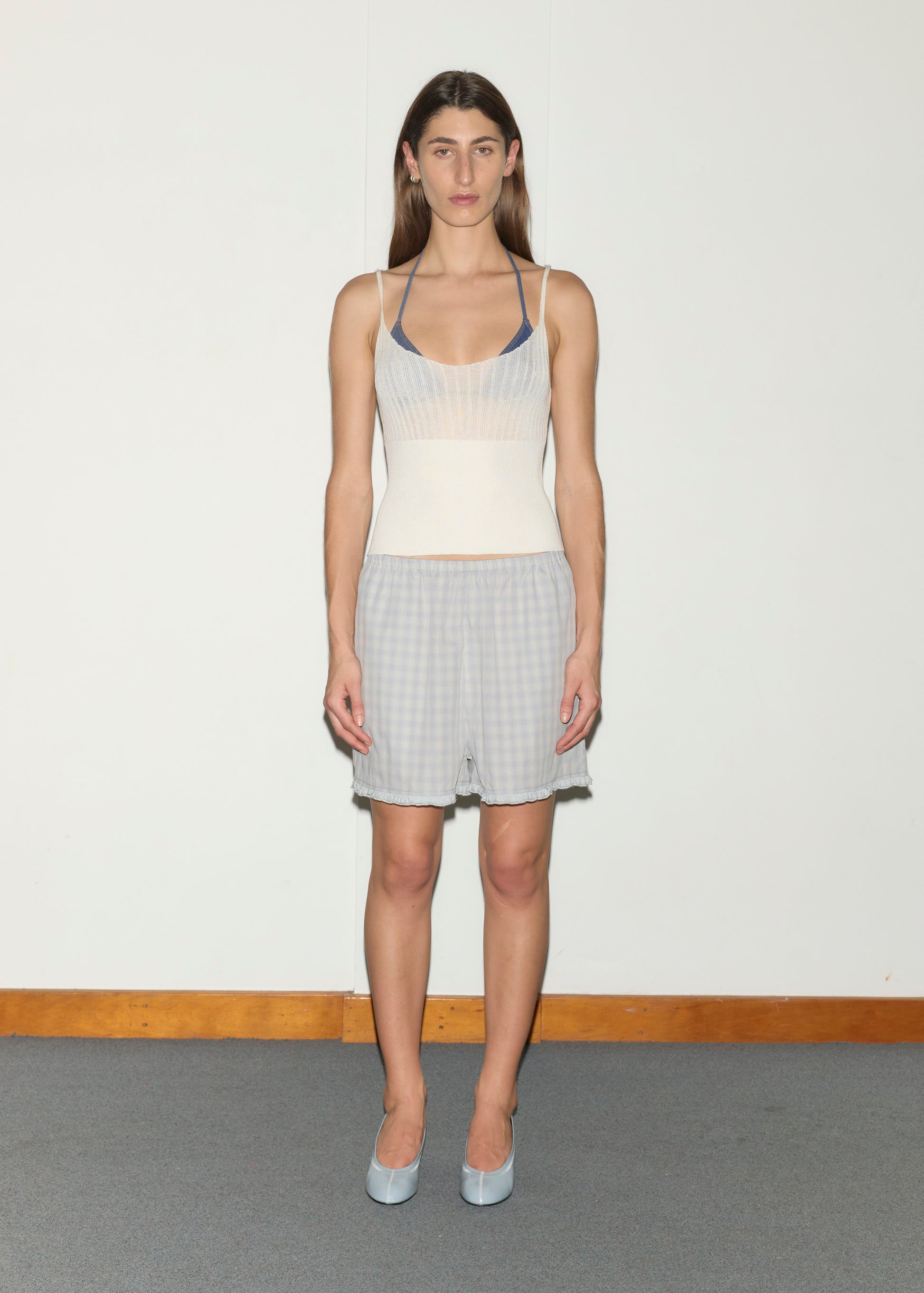A woman stands indoors on gray carpet against a white wall, wearing the Simple Short - Blue Check, a cream sleeveless top over a blue bralette, and light blue heels, looking directly at the camera.