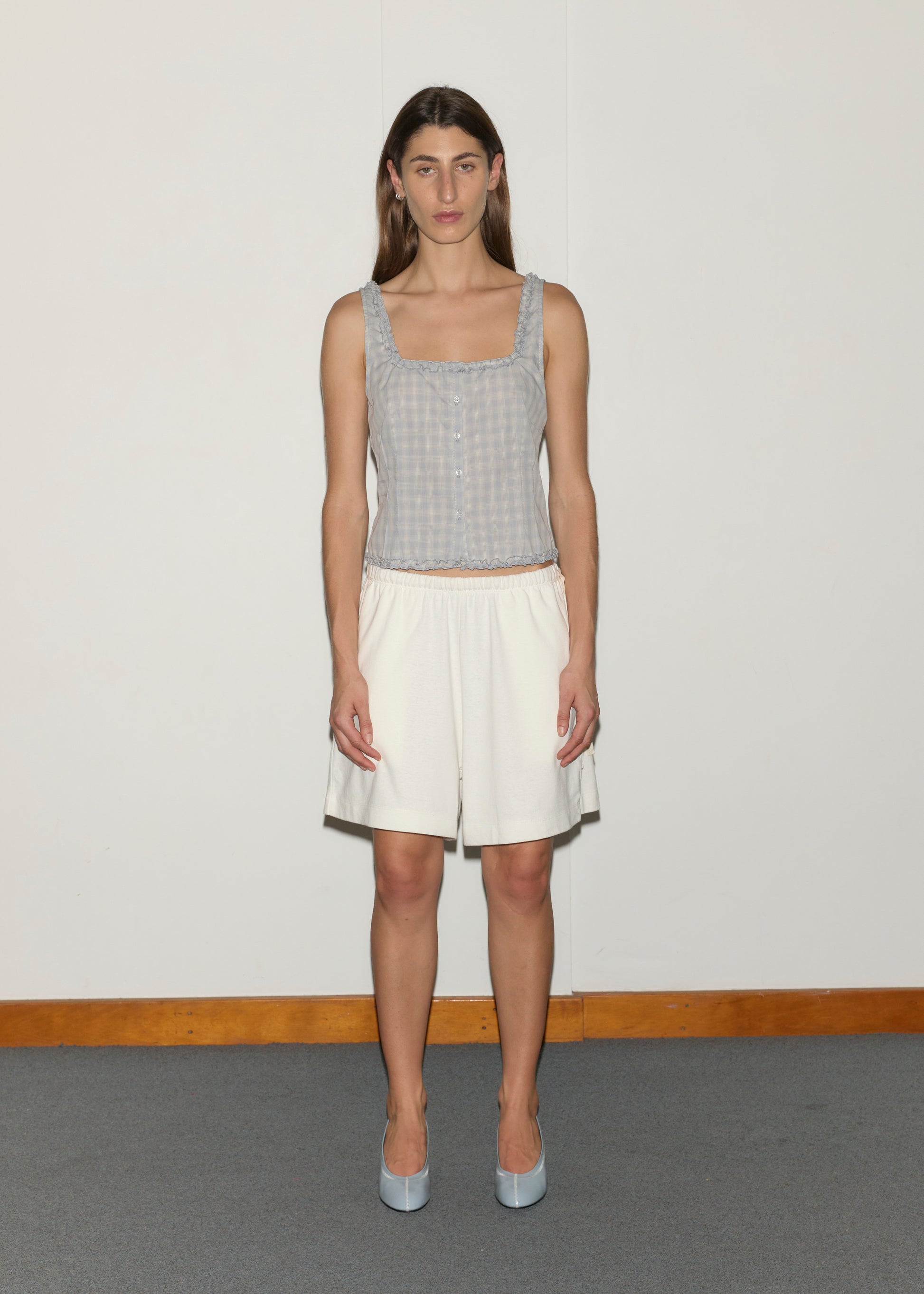 A person with long brown hair stands facing forward against a white wall, wearing a Deiji Studios light blue cotton poplin sleeveless top, Relax Short - Off White, blue shoes, with a wooden baseboard and gray floor in the background.