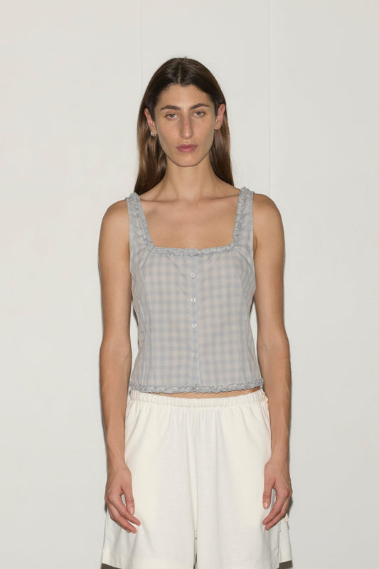 A woman with long brown hair and a neutral expression stands against a white wall, wearing the Button Frill Top - Blue Check by Deiji Studios, loose white shorts, and light blue heels, her arms relaxed at her sides.