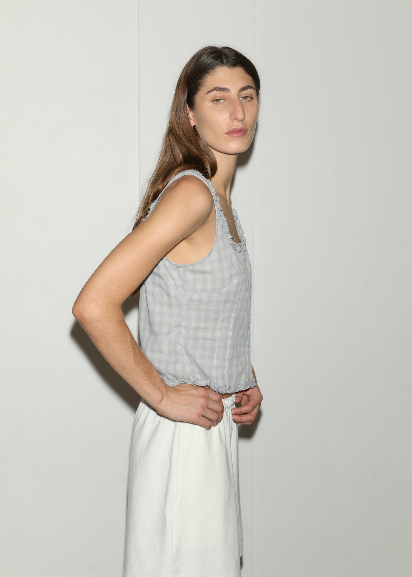 A person with long brown hair, wearing Deiji Studios’ Button Frill Top in blue check and a white skirt, stands sideways against a plain white wall, looking at the camera with a neutral expression.