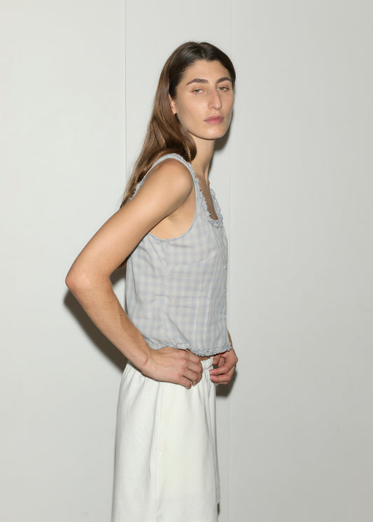 A person with long brown hair, wearing Deiji Studios’ Button Frill Top in blue check and a white skirt, stands sideways against a plain white wall, looking at the camera with a neutral expression.