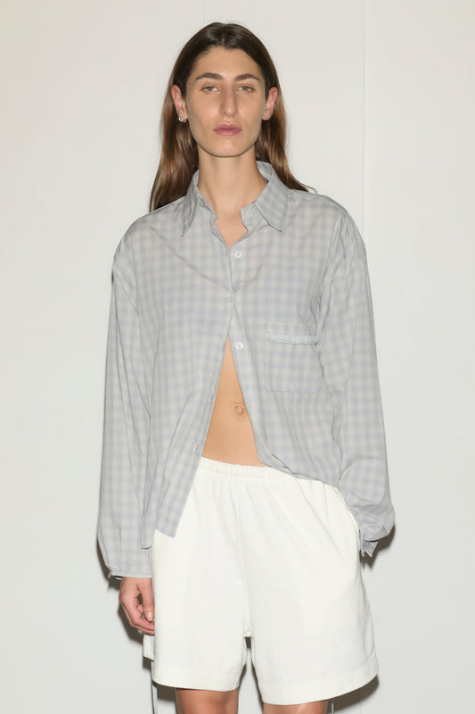 A person with long brown hair wears a light gray checked day shirt, partially unbuttoned, and loose white shorts, standing against a plain white background.