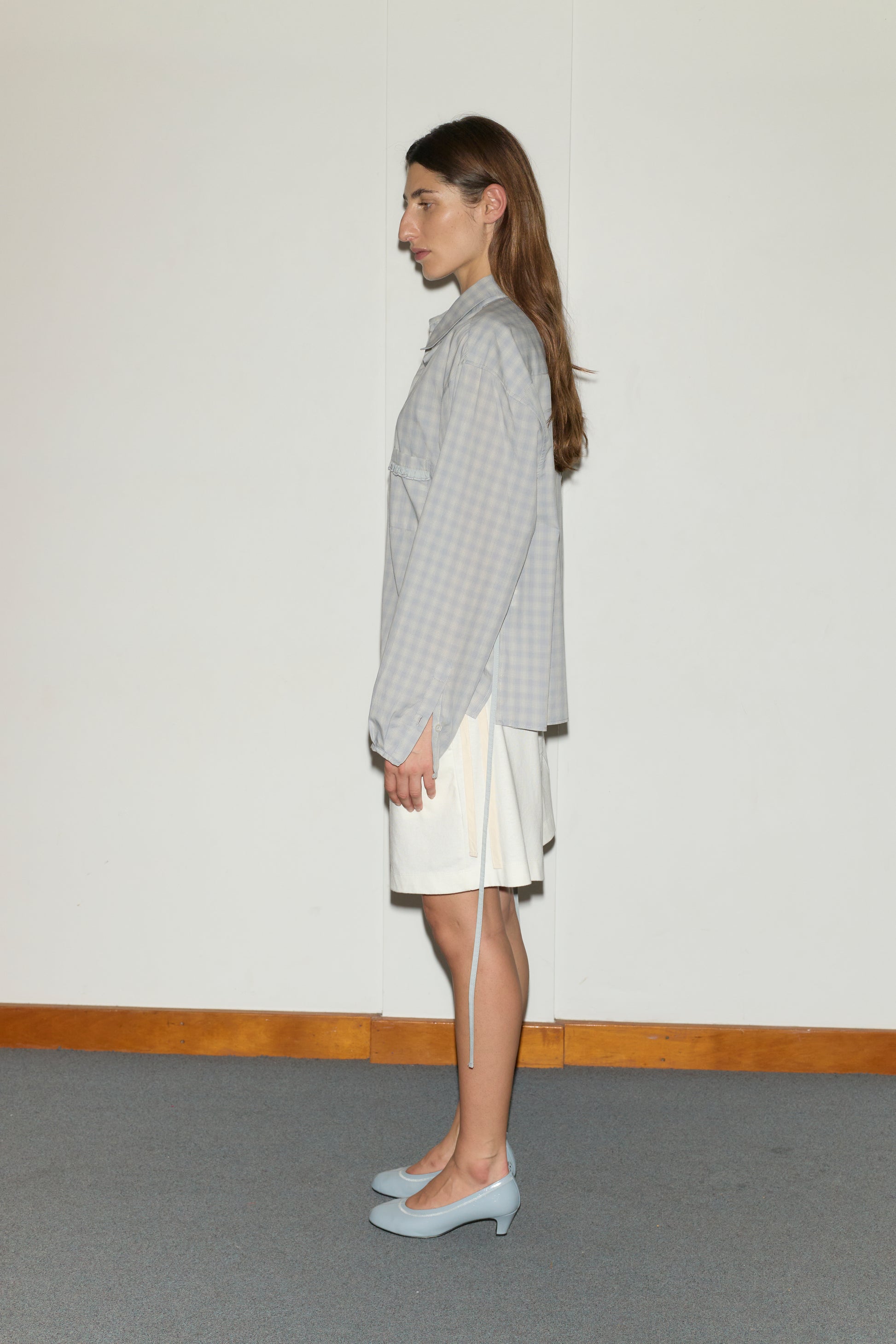 A woman with long brown hair stands in profile against a white wall, wearing the Day Shirt - Blue Check, a white skirt, and light blue heels on a carpeted floor edged with a wooden baseboard.
