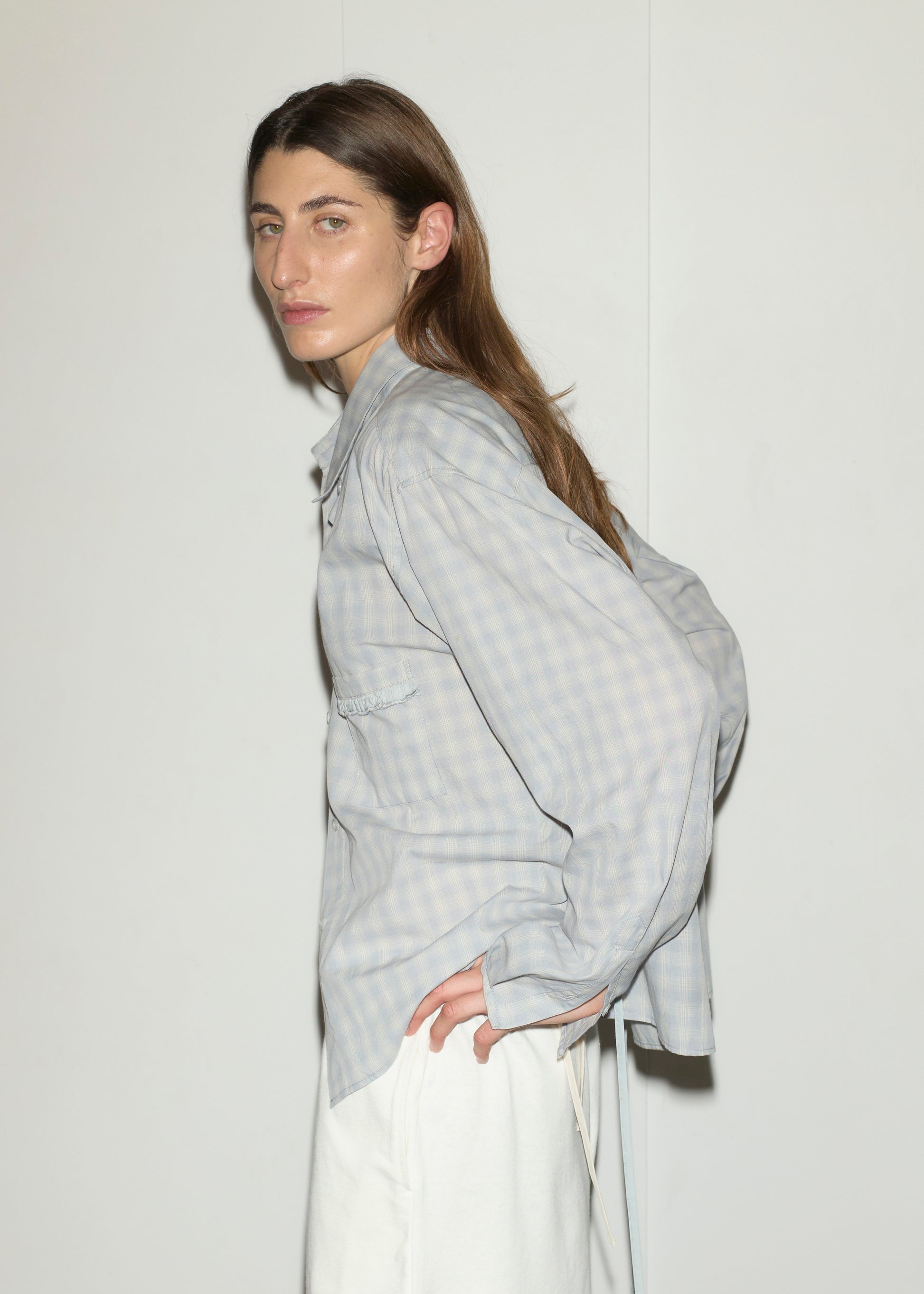 A person with long brown hair stands sideways against a white background, wearing the Day Shirt - Blue Check in a boxy fit with subtle blue checks and light grey tones, paired with white pants and one hand on their hip.