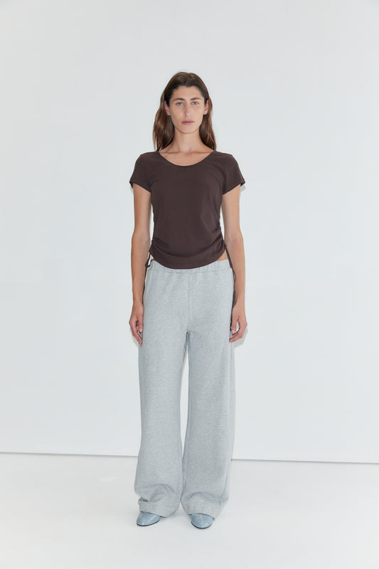 A woman stands against a plain white background, wearing the Draw Side Tee in brown—a relaxed cotton jersey short-sleeve top. She pairs it with loose light gray sweatpants and light blue shoes. Her straight brown hair frames her neutral expression.