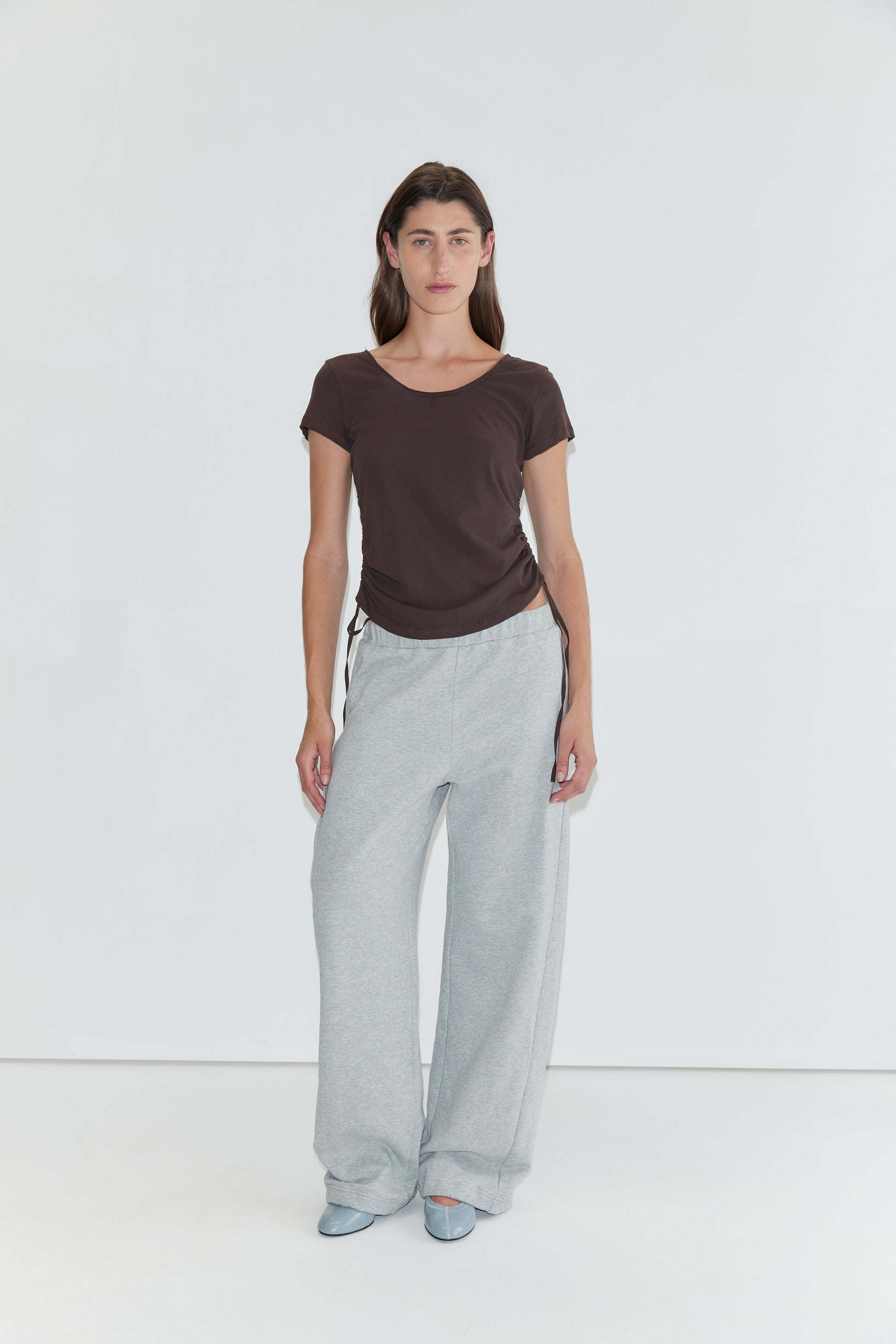 A person stands against a plain white background wearing a brown short-sleeved top, the Side Pleat Pant in Grey Marle—evoking Deiji Studios’ effortless style—and light blue shoes.