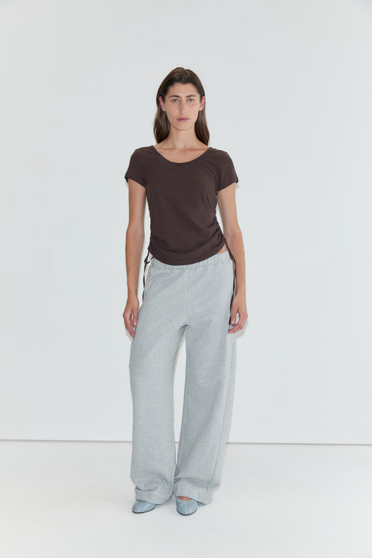 A person stands against a plain white background wearing a brown short-sleeved top, the Side Pleat Pant in Grey Marle—evoking Deiji Studios’ effortless style—and light blue shoes.