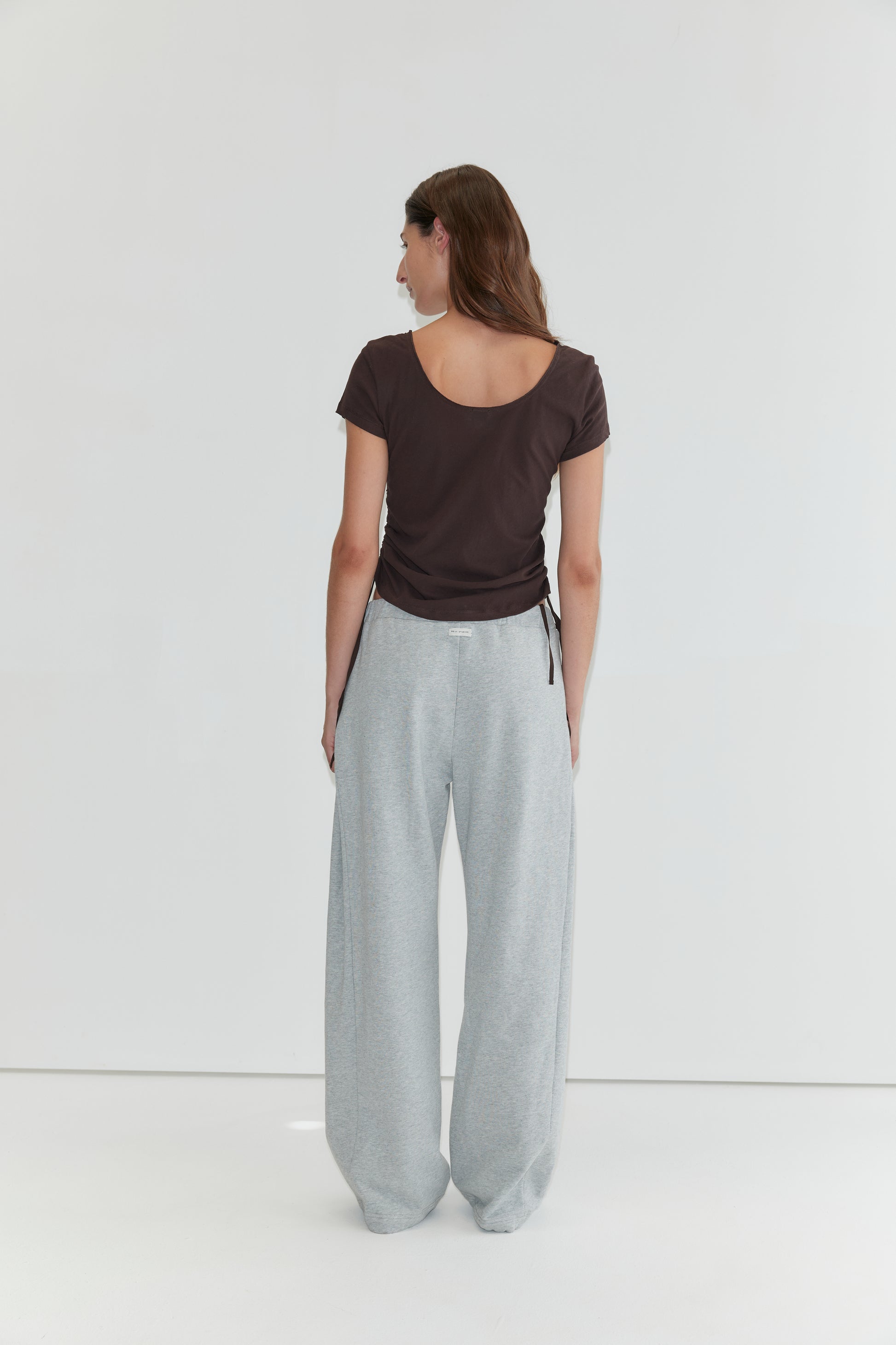 A person with long brown hair stands facing away, wearing the Draw Side Tee - Brown crafted from cotton jersey and loose light gray sweatpants, against a plain white background—a relaxed look reminiscent of deiji studios.