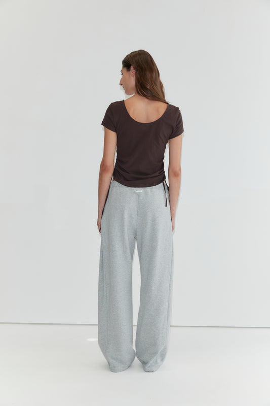 A person with long brown hair stands facing away, wearing the Draw Side Tee - Brown crafted from cotton jersey and loose light gray sweatpants, against a plain white background—a relaxed look reminiscent of deiji studios.